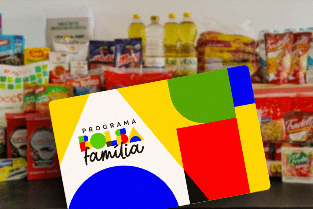 Cartão colorido do Programa Bolsa Família em primeiro plano. Ao fundo, borrados, estão diversos itens de supermercado, como óleo, arroz e pacotes de macarrão, sugerindo uma cesta básica.