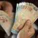 Notas de 50 reais em mãos, representando o 13º salário para brasileiros." Título: "Governo anuncia 13º salário para brasileiros