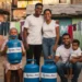 Família sorrindo em uma rua, com botijões de gás azuis do programa Gás do Povo. Uma senhora segura um botijão, enquanto a família se posiciona ao lado.