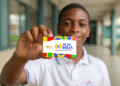 Jovem estudante negro do ensino médio segurando, em destaque, o cartão do programa "Pé-de-Meia" com as cores da bandeira do Brasil. O rapaz sorri ao fundo, em um corredor escolar.