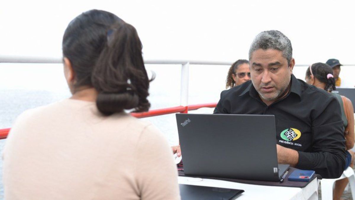 Servidor do INSS com camiseta da Previdência Social atendendo uma segurada em um notebook durante mutirão de atendimento.