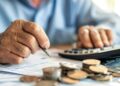 Idoso calcula aposentadoria com moedas, papéis e calculadora sobre a mesa.