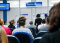 Sala de espera movimentada em posto do INSS para perícia médica presencial, com painel de chamada de senhas.