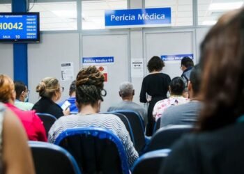 Sala de espera movimentada em posto do INSS para perícia médica presencial, com painel de chamada de senhas.