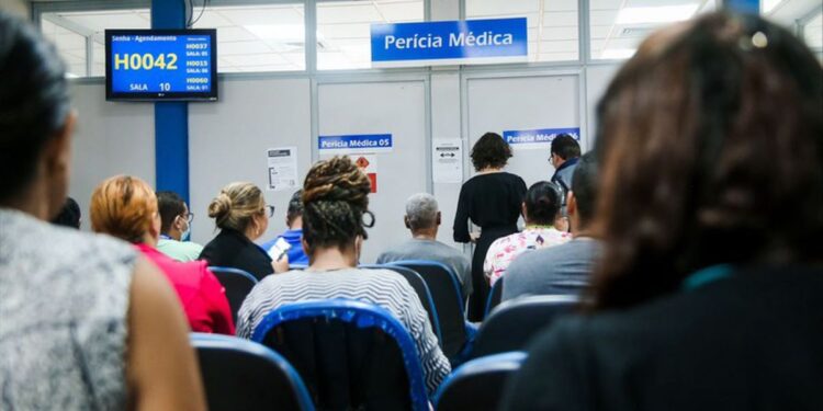 Sala de espera movimentada em posto do INSS para perícia médica presencial, com painel de chamada de senhas.