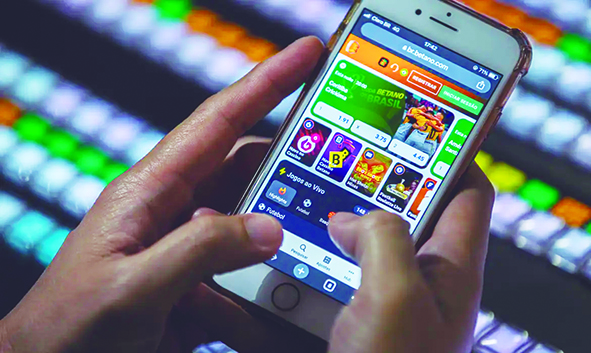 Uma pessoa segura um smartphone com o aplicativo de uma plataforma de apostas esportivas aberta na tela. Os dedos estão posicionados para interagir com o site, que mostra opções de jogos e apostas ao vivo.