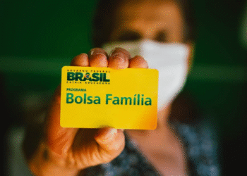 Cartão amarelo do programa Bolsa Família em destaque, com a pessoa ao fundo usando máscara.