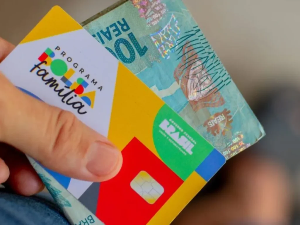 Cartão do Bolsa Família sendo segurado junto com cédula de cem reais, simbolizando pagamento do benefício