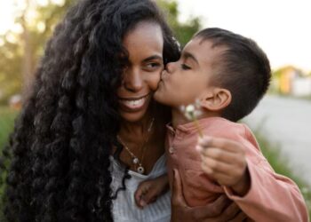 Criança dá beijo carinhoso na mãe sorridente em momento especial ao ar livre.