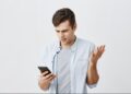 Homem jovem com expressão confusa e gesticulando com a mão, olhando para o celular, com fundo branco. Este texto é crucial para a acessibilidade, descrevendo a imagem para pessoas com deficiência visual e ajudando o Google a entender o conteúdo visual.
