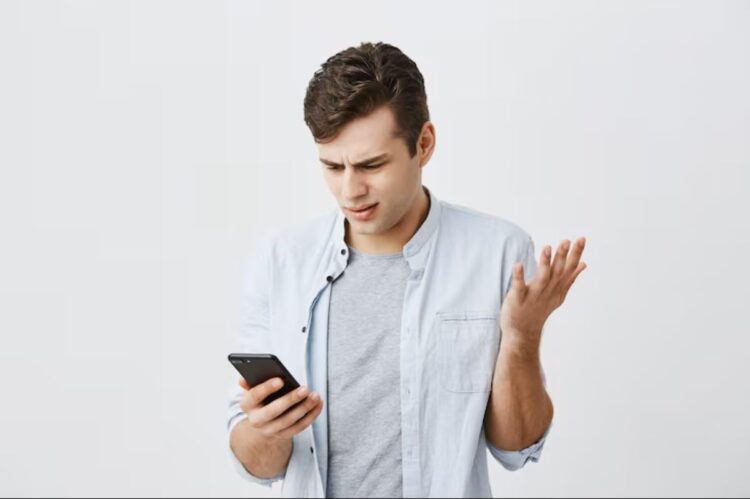 Homem jovem com expressão confusa e gesticulando com a mão, olhando para o celular, com fundo branco. Este texto é crucial para a acessibilidade, descrevendo a imagem para pessoas com deficiência visual e ajudando o Google a entender o conteúdo visual.