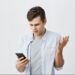 Homem jovem com expressão confusa e gesticulando com a mão, olhando para o celular, com fundo branco. Este texto é crucial para a acessibilidade, descrevendo a imagem para pessoas com deficiência visual e ajudando o Google a entender o conteúdo visual.