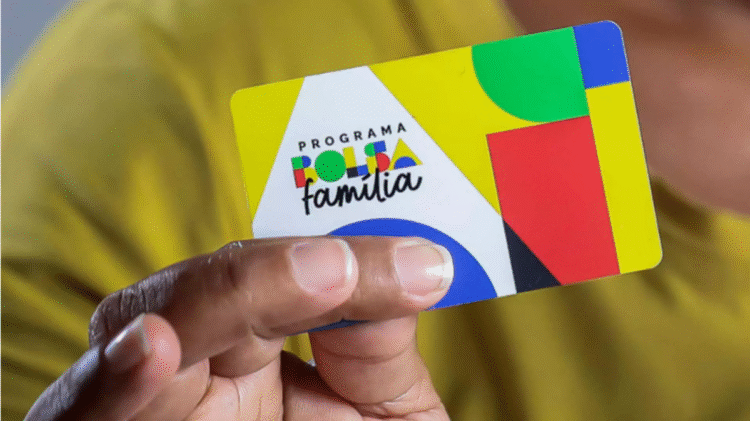 Pessoa segurando o novo cartão do Bolsa Família sobre fundo amarelo.
