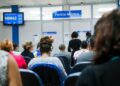 Pessoas aguardando atendimento na fila do INSS para a Perícia Médica, com o painel eletrônico de senhas exibindo a chamada 'H0042'.