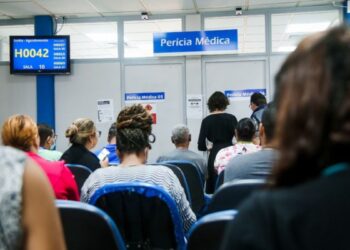 Pessoas aguardando atendimento na fila do INSS para a Perícia Médica, com o painel eletrônico de senhas exibindo a chamada 'H0042'.