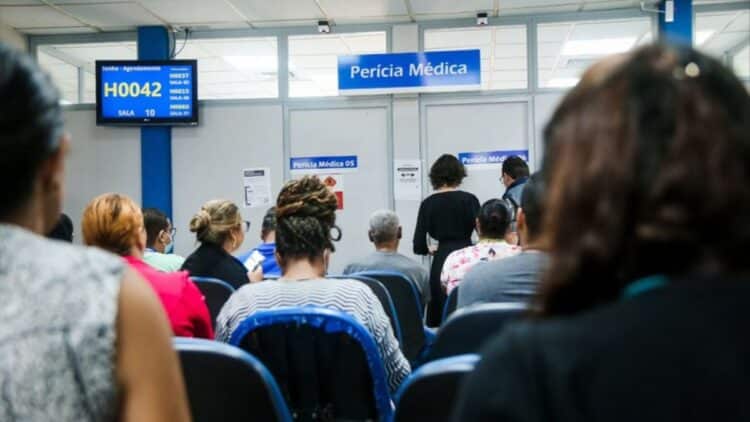Pessoas aguardando atendimento na fila do INSS para a Perícia Médica, com o painel eletrônico de senhas exibindo a chamada 'H0042'.