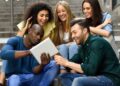 Grupo de jovens estudantes diversos e sorrindo, interagindo com um tablet em um ambiente urbano.