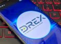 Tela de um celular mostrando o logo da moeda digital Drex do Banco Central do Brasil, com um teclado de computador ao fundo.