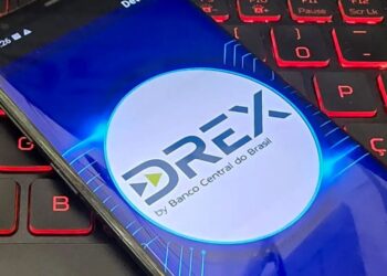 Tela de um celular mostrando o logo da moeda digital Drex do Banco Central do Brasil, com um teclado de computador ao fundo.