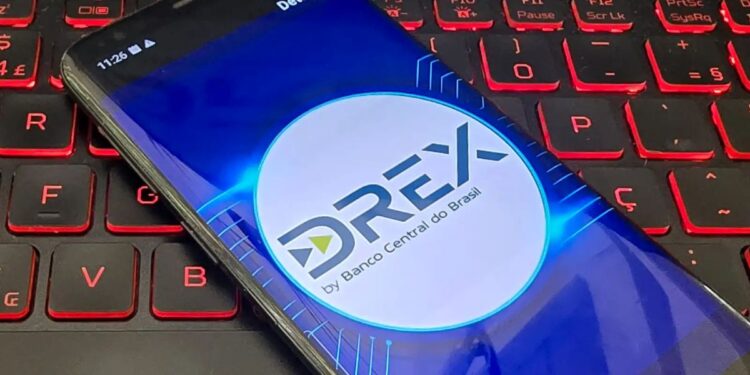 Tela de um celular mostrando o logo da moeda digital Drex do Banco Central do Brasil, com um teclado de computador ao fundo.