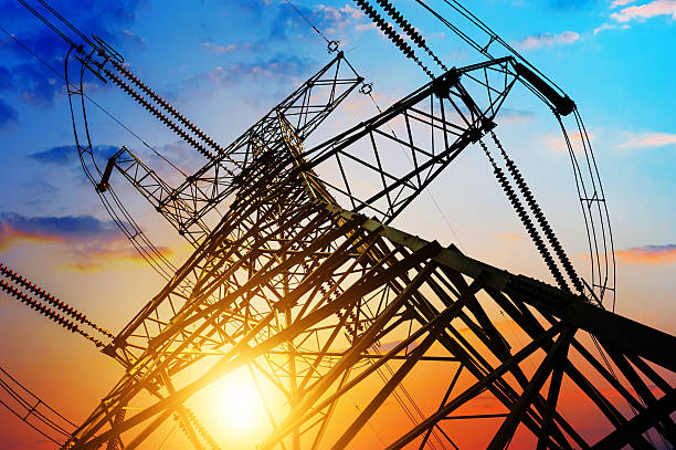 Uma torre de transmissão de energia elétrica em silhueta contra um céu colorido de fim de tarde, com o sol se pondo no horizonte, referenciando a nova tarifa social de energia que dará gratuidade as famílias mais necessitadas. 