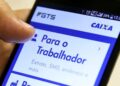 Mão de uma pessoa segurando um smartphone com o aplicativo do FGTS da Caixa aberto na tela. A seção 'Para o Trabalhador' está em destaque, mostrando opções como 'Extrato, SMS, endereço e mais', fazendo referência ao saque-aniversário do FGTS.