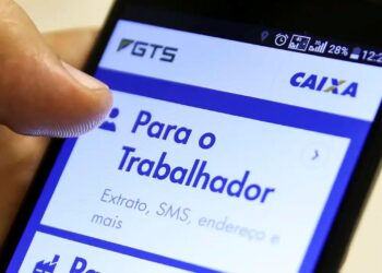 Mão de uma pessoa segurando um smartphone com o aplicativo do FGTS da Caixa aberto na tela. A seção 'Para o Trabalhador' está em destaque, mostrando opções como 'Extrato, SMS, endereço e mais', fazendo referência ao saque-aniversário do FGTS.