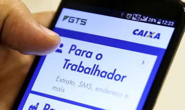 Mão de uma pessoa segurando um smartphone com o aplicativo do FGTS da Caixa aberto na tela. A seção 'Para o Trabalhador' está em destaque, mostrando opções como 'Extrato, SMS, endereço e mais', fazendo referência ao saque-aniversário do FGTS.