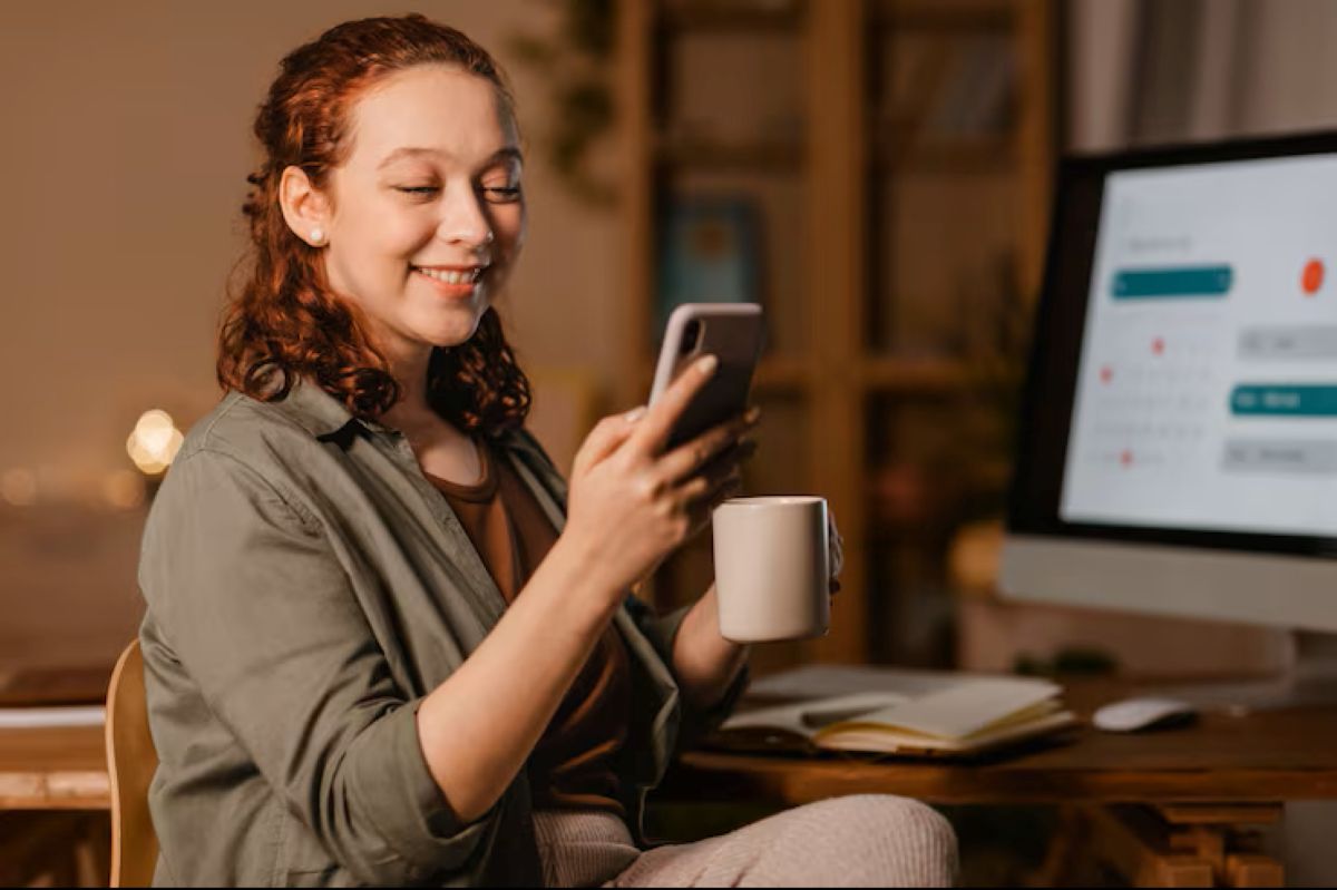 Freelancer ruiva em home office noturno, sorrindo ao usar o celular e segurar uma caneca, ilustrando a flexibilidade de horário e a gestão de comunicação para o sucesso no trabalho sob demanda.