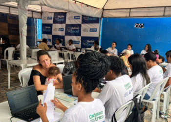 Mutirão do CadÚnico com pessoas em mesas sob tenda, usando camisetas brancas, em atividade de cadastro.
