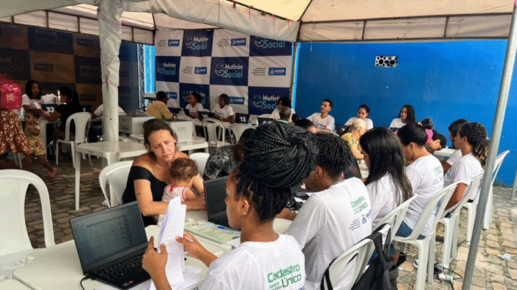 Mutirão do CadÚnico com pessoas em mesas sob tenda, usando camisetas brancas, em atividade de cadastro.