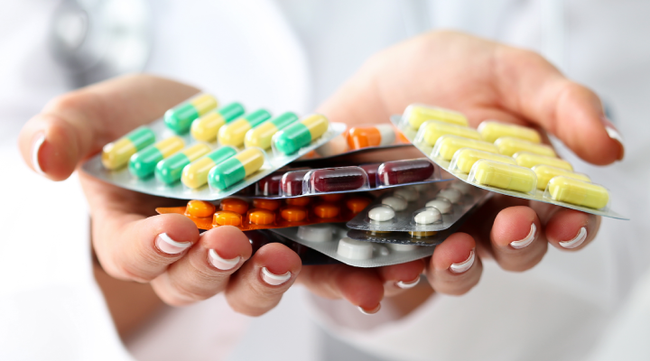 Medicamentos variados em blister sendo segurados por mãos de uma pessoa com jaleco. A imagem ilustra a variedade de remédios que podem ser disponibilizados em farmácias dentro de supermercados.