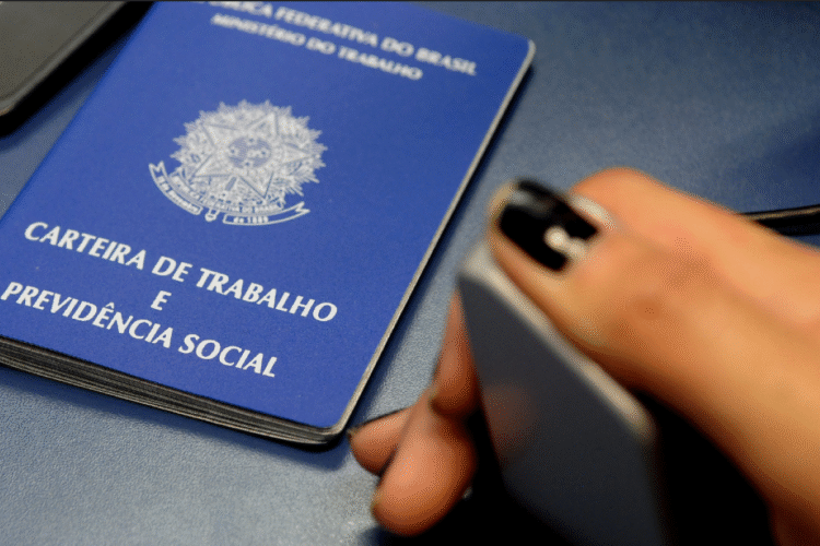 Carteira de Trabalho e Previdência Social azul sobre uma mesa, com uma mão segurando um celular.