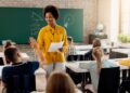Uma professora negra e sorridente, vestindo uma blusa amarela, conversa com um aluno em uma sala de aula.