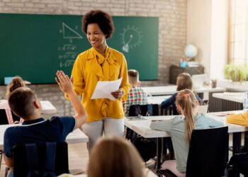 Uma professora negra e sorridente, vestindo uma blusa amarela, conversa com um aluno em uma sala de aula.