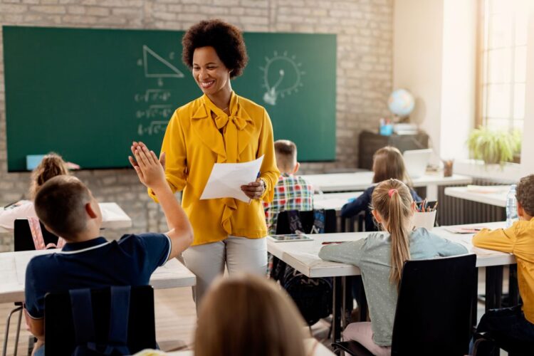 Uma professora negra e sorridente, vestindo uma blusa amarela, conversa com um aluno em uma sala de aula.