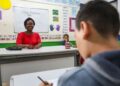 Professora sorridente em sala de aula durante aula com aluno ao fundo