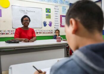 Professora sorridente em sala de aula durante aula com aluno ao fundo