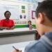 Professora sorridente em sala de aula durante aula com aluno ao fundo