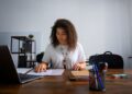 Mulher sorridente e concentrada trabalhando com documentos em home office, usando notebook e mesa de madeira. Representa a flexibilidade e o foco do trabalho autônomo e a busca por jobs sob demanda em plataformas digitais.