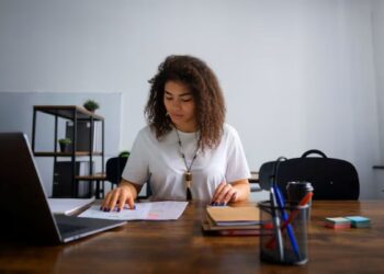 Mulher sorridente e concentrada trabalhando com documentos em home office, usando notebook e mesa de madeira. Representa a flexibilidade e o foco do trabalho autônomo e a busca por jobs sob demanda em plataformas digitais.