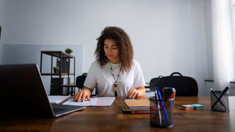 Mulher sorridente e concentrada trabalhando com documentos em home office, usando notebook e mesa de madeira. Representa a flexibilidade e o foco do trabalho autônomo e a busca por jobs sob demanda em plataformas digitais.