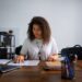 Mulher sorridente e concentrada trabalhando com documentos em home office, usando notebook e mesa de madeira. Representa a flexibilidade e o foco do trabalho autônomo e a busca por jobs sob demanda em plataformas digitais.