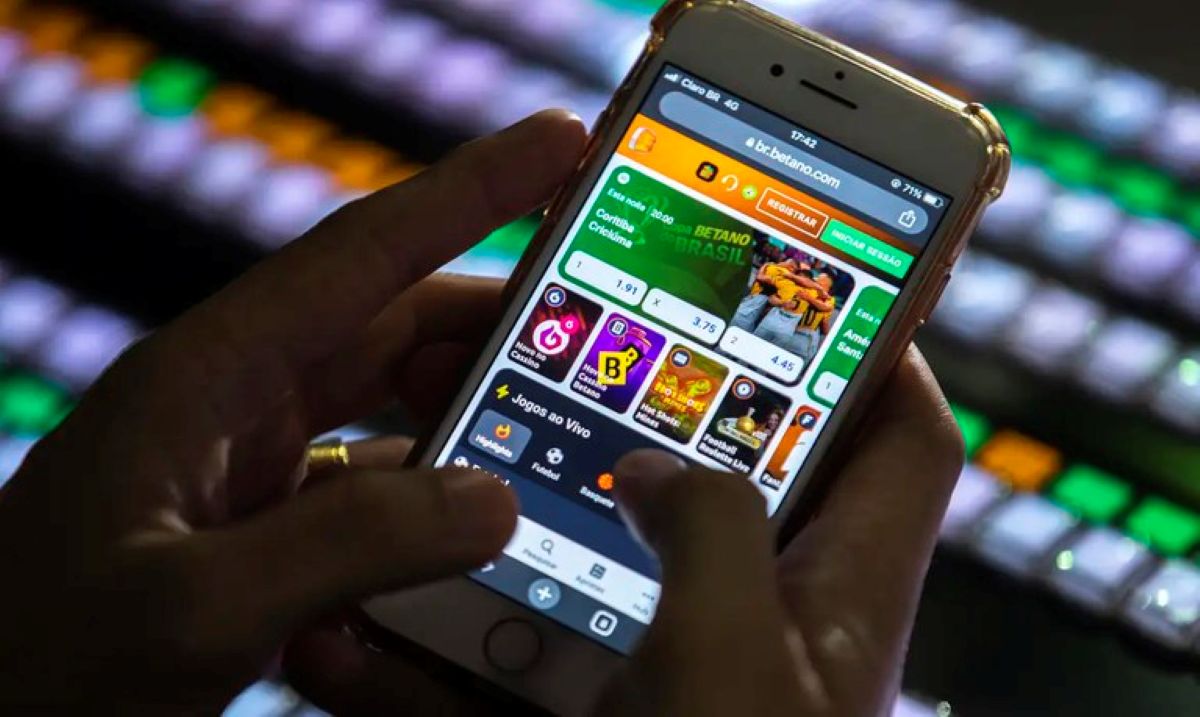 Mãos de pessoa utilizando celular com o aplicativo de uma casa de apostas esportivas (BET) aberta, ilustrando o bloqueio de depósitos para beneficiários do Bolsa Família e BPC.