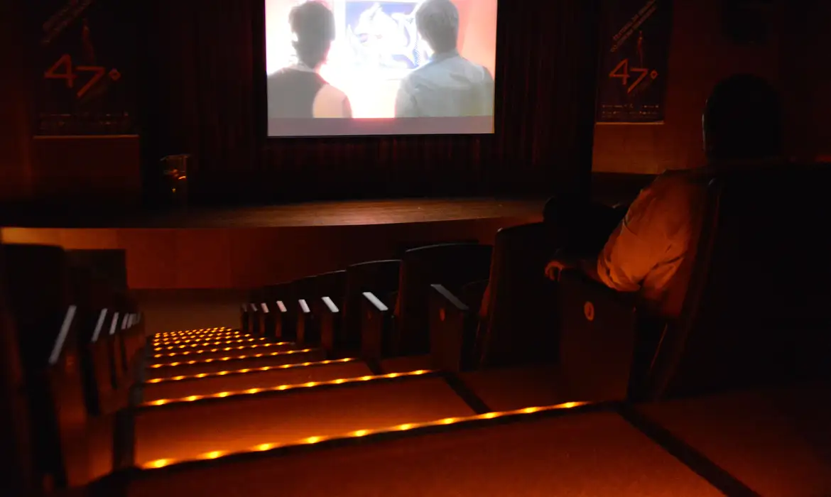 Interior de um auditório ou sala de cinema escurecida com poltronas vazias e iluminação de segurança nas escadas, representando o local onde o documento de meia-entrada (como o ID Jovem ou carteira de estudante) deve ser apresentado.