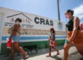 Pessoas, incluindo uma criança, caminhando em frente a um prédio com o letreiro 'CRAS - Centro de Referência de Assistência Social', indicando a busca por serviços sociais.