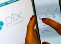 Mão feminina segurando um smartphone com a tela do aplicativo Pix aberta, mostrando opções de pagamento e leitura de QR Code, com a logo Pix ao fundo, ilustrando a função Pix por foto.