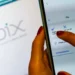 Mão feminina segurando um smartphone com a tela do aplicativo Pix aberta, mostrando opções de pagamento e leitura de QR Code, com a logo Pix ao fundo, ilustrando a função Pix por foto.