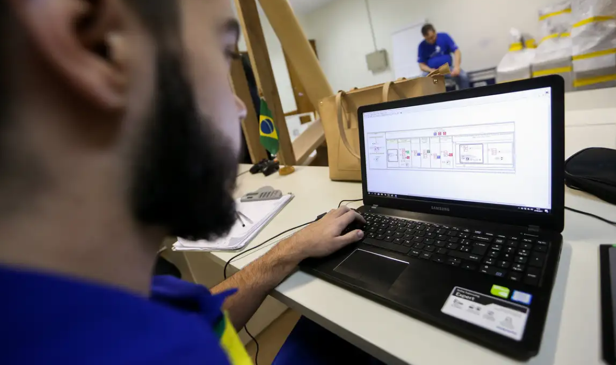Um homem com barba, vestindo uma camisa azul com detalhes amarelos, trabalhando em um laptop Samsung. A tela exibe um esquema digital. Ao fundo, uma bandeira do Brasil e outro profissional borrado.