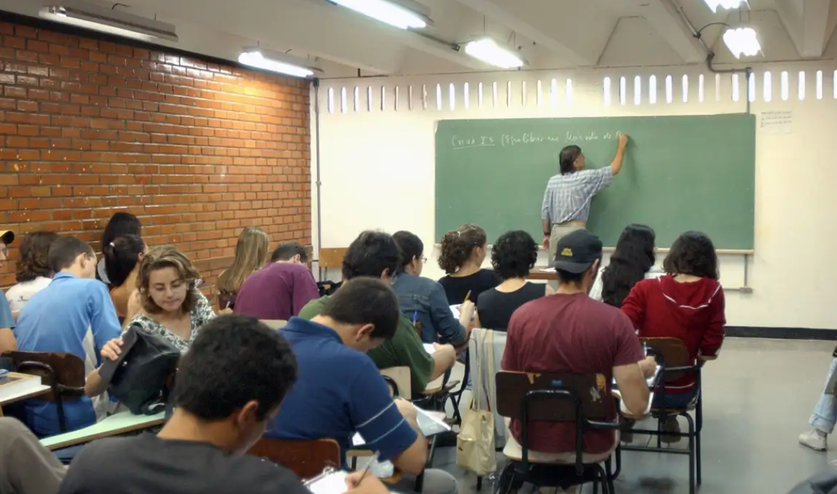Professor escrevendo em quadro-negro para alunos em sala de aula, ilustrando o ambiente de trabalho em dia útil